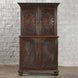 The Bhuv Teak 2 Part Cabinet by John Robshaw features ornate carved doors with circular motifs and decorative trim, displayed against a white textured brick wall on a concrete floor. Showcasing exquisite carved detailing throughout, this piece exemplifies vintage furniture craftsmanship.