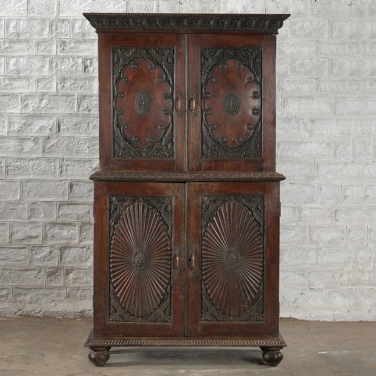 The Bhuv Teak 2 Part Cabinet by John Robshaw features ornate carved doors with circular motifs and decorative trim, displayed against a white textured brick wall on a concrete floor. Showcasing exquisite carved detailing throughout, this piece exemplifies vintage furniture craftsmanship.