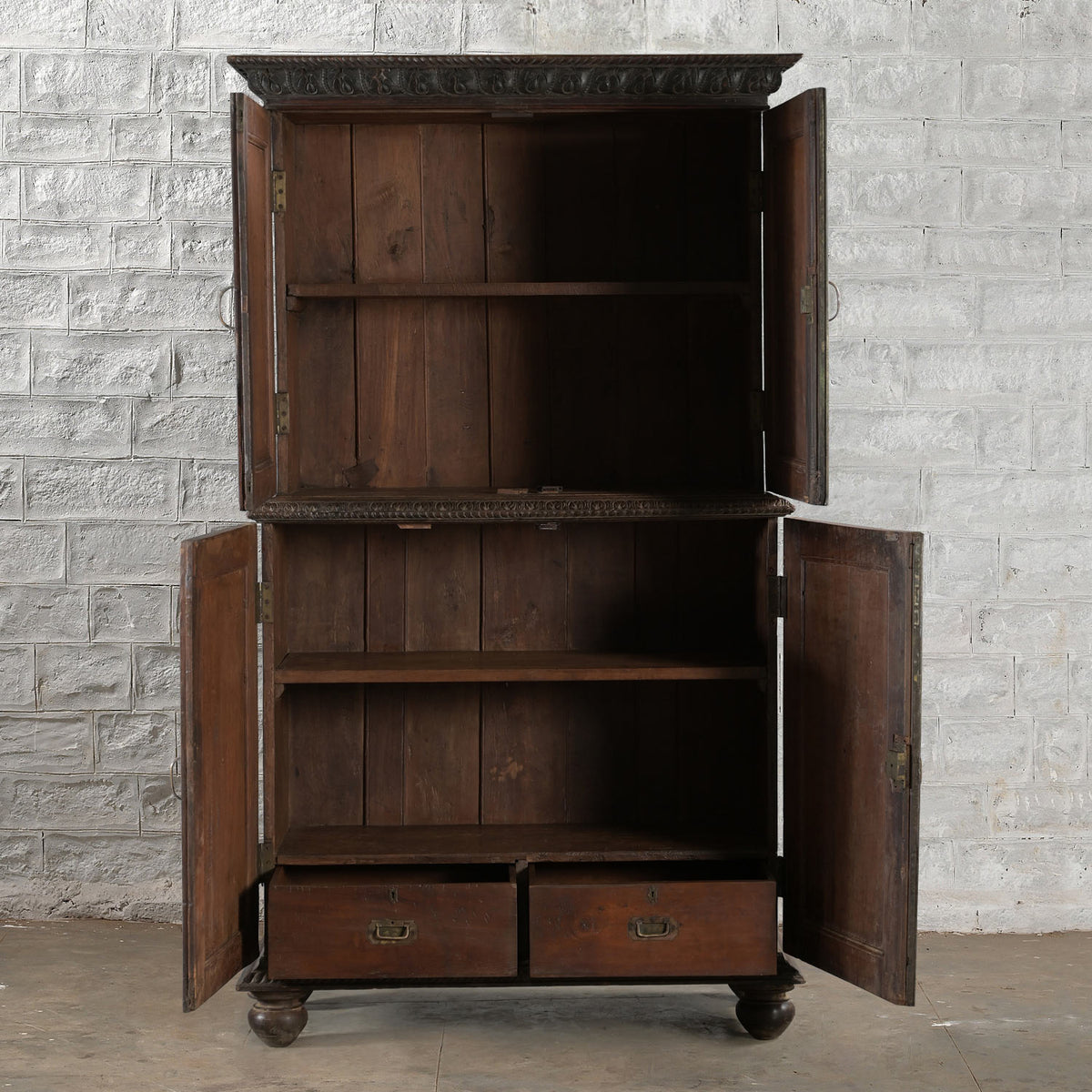The Bhuv Teak 2 Part Cabinet by John Robshaw stands against a white brick wall, featuring four open doors that reveal two shelves in the top section, one shelf in the bottom section, and two pull-out drawers at the base. Subtle carved details add to its timeless charm, making this vintage-inspired piece a standout addition to any space.