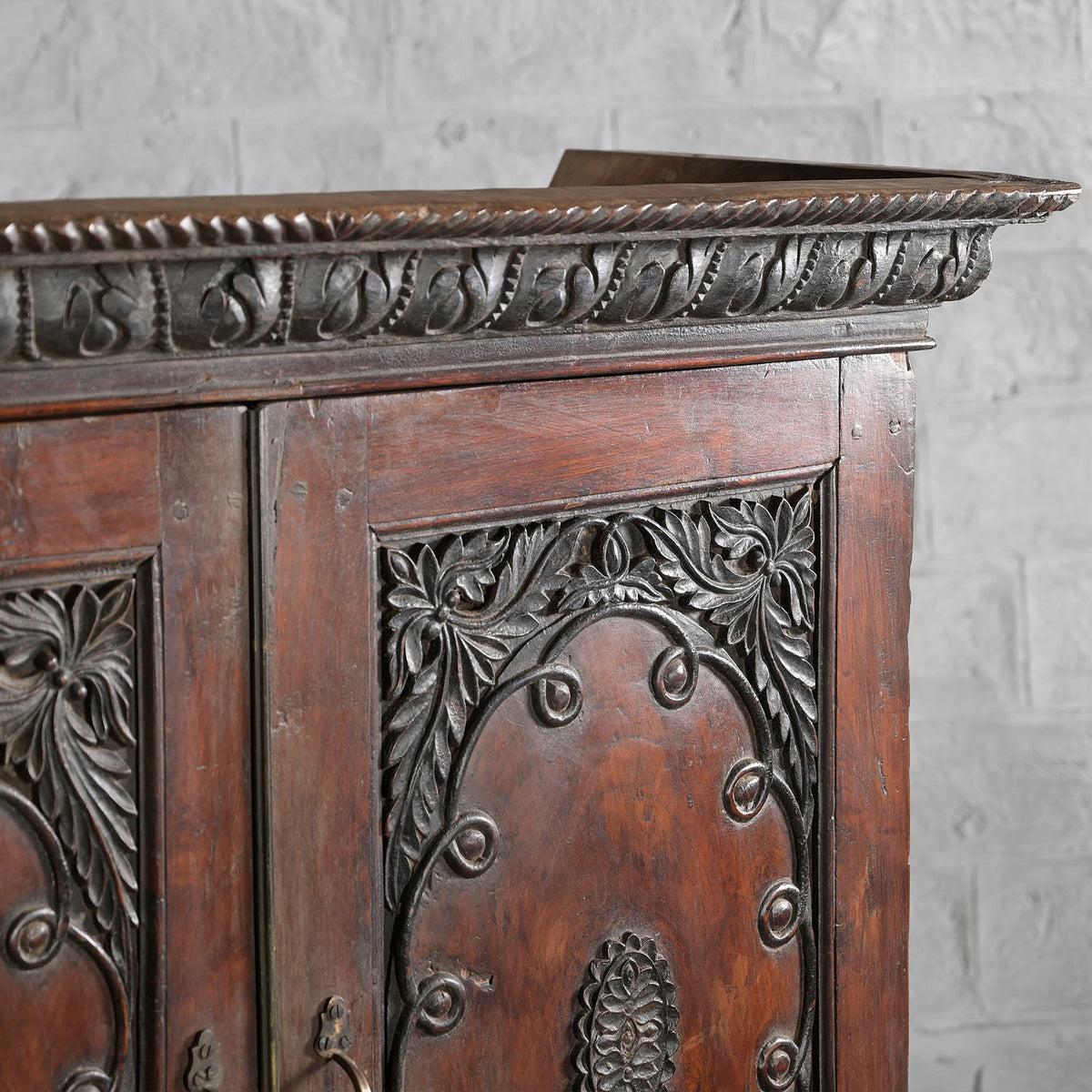 Close-up of the John Robshaw Bhuv Teak 2 Part Cabinet, featuring intricately carved floral and leaf patterns in solid teak, set against a white brick wall background.