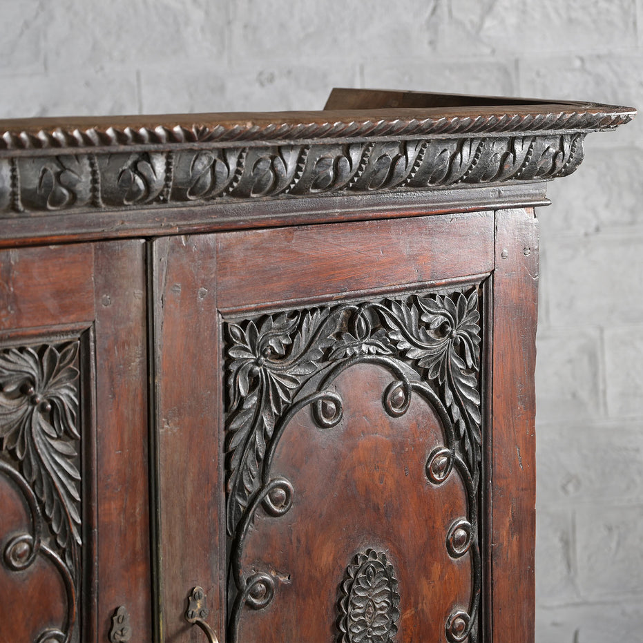Close-up of the John Robshaw Bhuv Teak 2 Part Cabinet, featuring intricately carved floral and leaf patterns in solid teak, set against a white brick wall background.