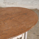 The John Robshaw Teak Table 10 features a round, handcrafted teak design with a weathered brown top and visible wood grain. Set against a concrete floor and light brick wall, this table is perfect for those seeking vintage furniture charm.