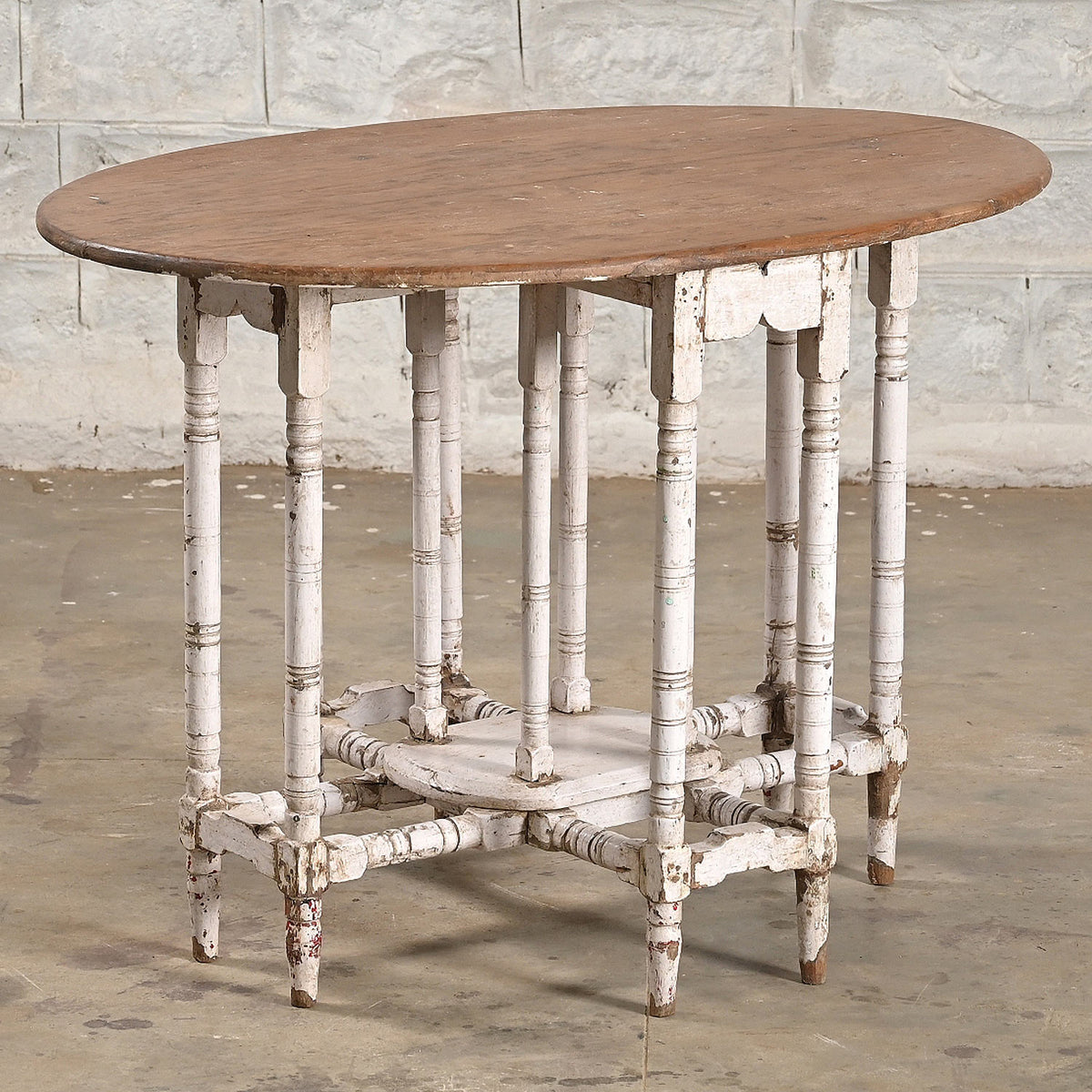 John Robshaw’s Teak Table 10 features an oval teak wood top with a weathered brown finish and distressed white spindle legs, set against a gray stone wall and concrete floor—a beautiful example of vintage furniture crafted in timeless style.