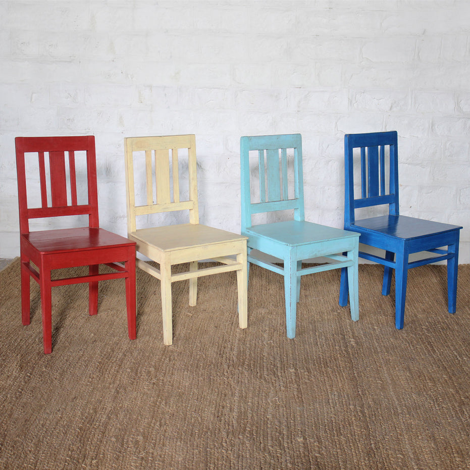 Four Chitrit Indigo School Chairs by John Robshaw, in red, yellow, light blue, and blue—each a classic school chair—are arranged in a row on a brown woven rug against a white brick wall.