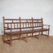 The Shahad Teak Bench by John Robshaw, showcasing a vintage design with a honey-toned finish, slatted seat and backrest, four sturdy legs, and two armrests, is positioned on a woven rug against a white brick wall.