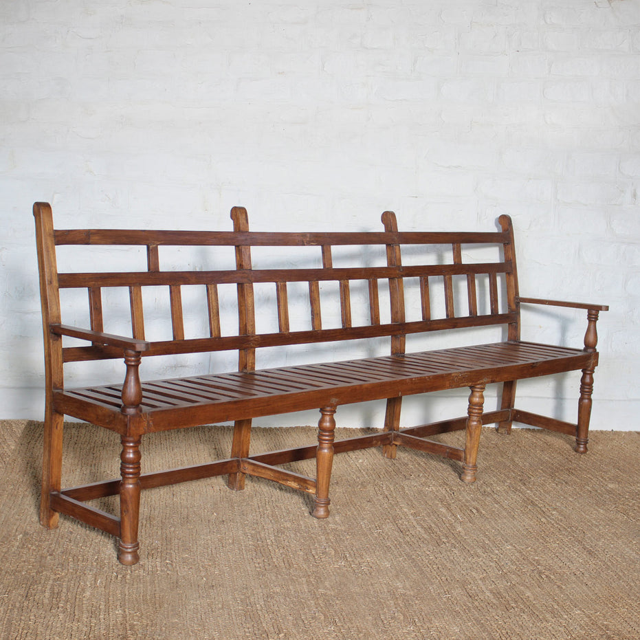 The Shahad Teak Bench by John Robshaw, showcasing a vintage design with a honey-toned finish, slatted seat and backrest, four sturdy legs, and two armrests, is positioned on a woven rug against a white brick wall.