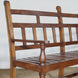The Shahad Teak Bench by John Robshaw features a vintage design with a slatted seat and turned legs, highlighted by vertical and horizontal slats on the backrest and armrests. It’s shown here set against a white wall atop a woven floor mat.