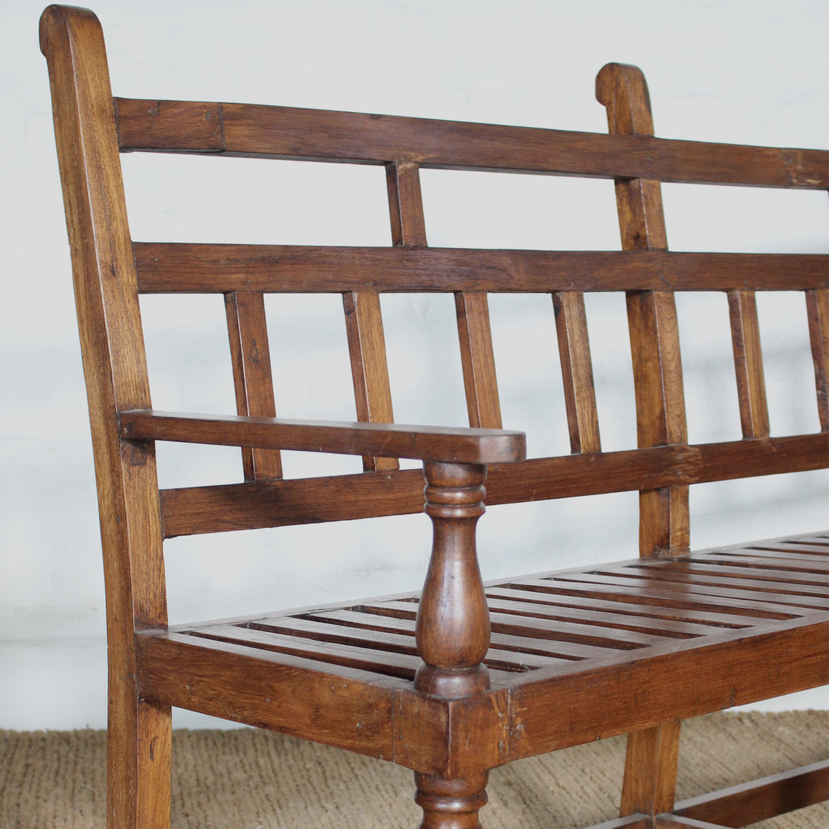 The Shahad Teak Bench by John Robshaw features a vintage design with a slatted seat and turned legs, highlighted by vertical and horizontal slats on the backrest and armrests. It’s shown here set against a white wall atop a woven floor mat.