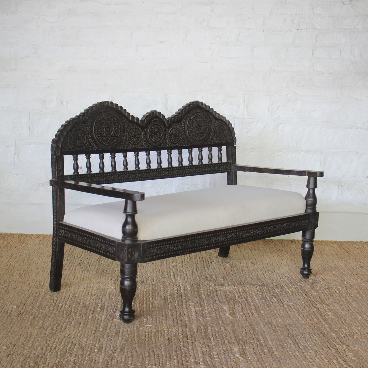 The Himavat Teak Seating Set by John Robshaw features ornately hand-carved backrests and arms in dark wood, with a light upholstered seat, displayed on a woven rug against a white brick wall.