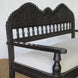 The Himavat Teak Seating Set by John Robshaw features a dark wood bench with a hand-carved backrest and armrests, detailed joinery, and a white upholstered seat, elegantly placed on a woven mat against a white brick wall.