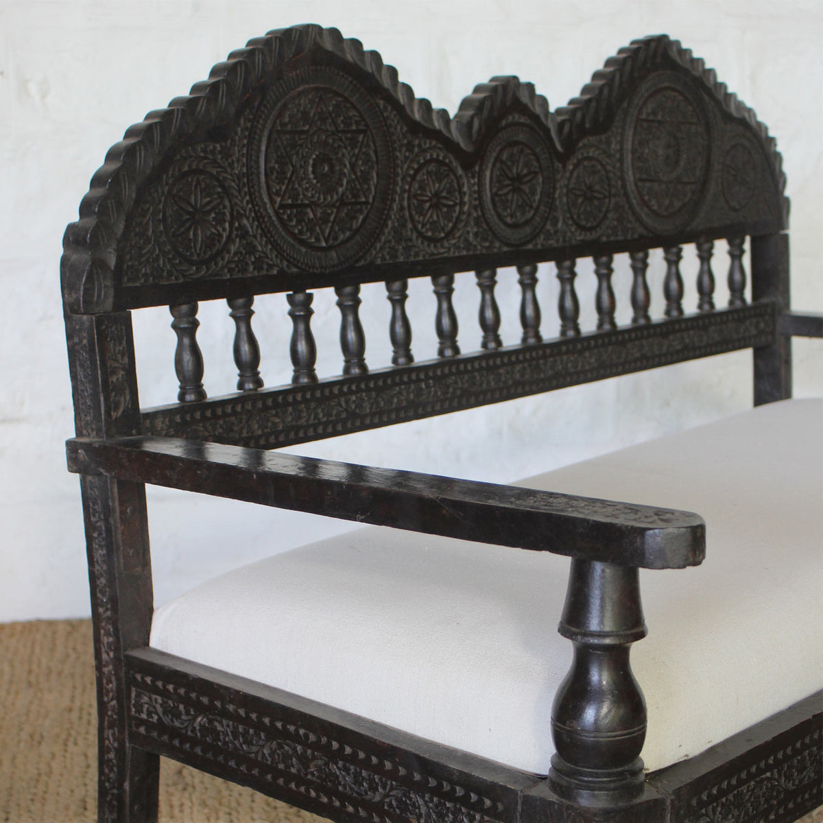 The Himavat Teak Seating Set by John Robshaw features a dark wood bench with a hand-carved backrest and armrests, detailed joinery, and a white upholstered seat, elegantly placed on a woven mat against a white brick wall.