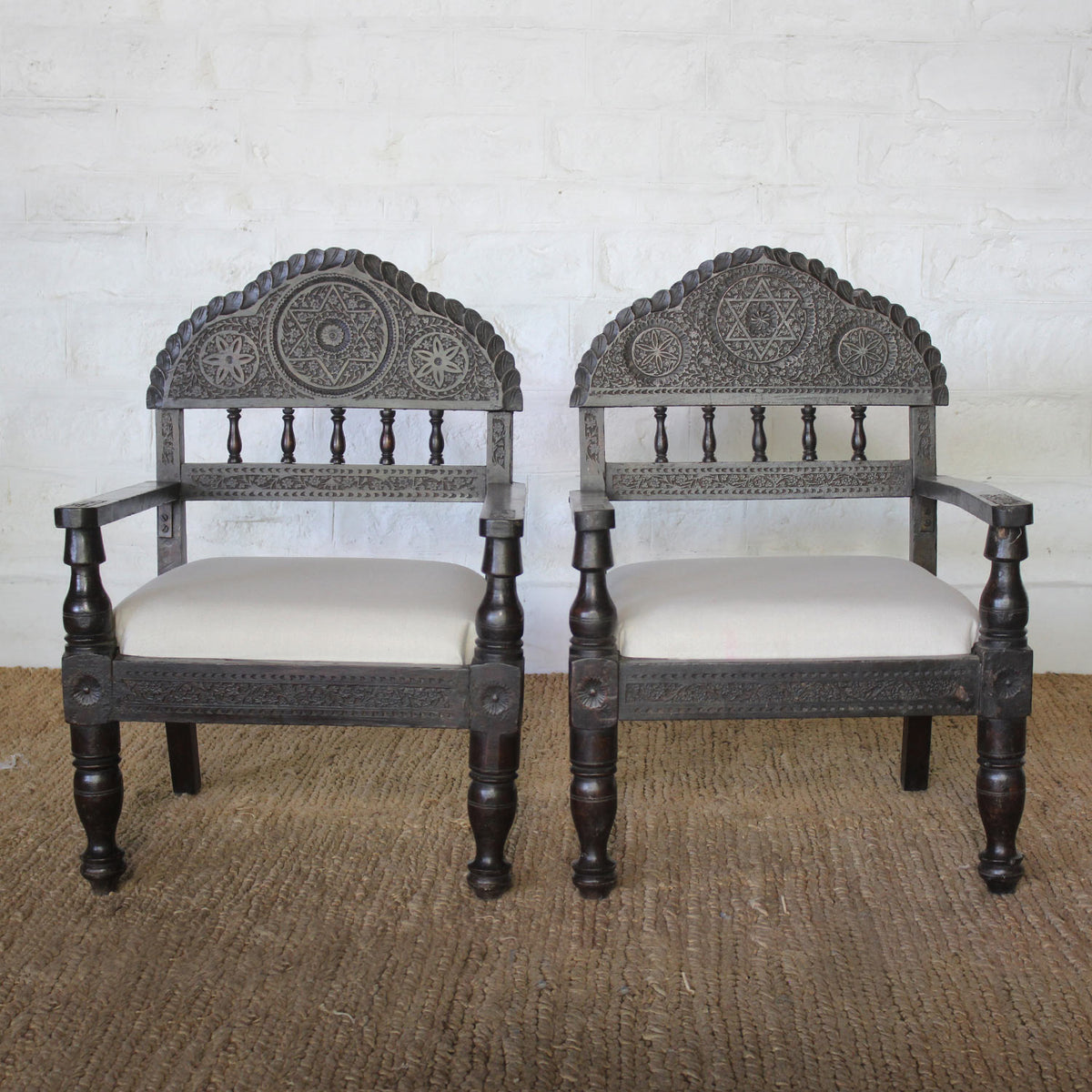 The Himavat Teak Seating Set by John Robshaw features two wooden armchairs with hand-carved backrests and detailed joinery, upholstered in light-colored fabric, placed side by side on a woven rug against a white brick wall.