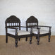 The Himavat Teak Seating Set by John Robshaw, featuring two dark wood armchairs with detailed joinery, hand-carved backrests, and white upholstered seats, is arranged side by side on a woven rug against a white brick wall.