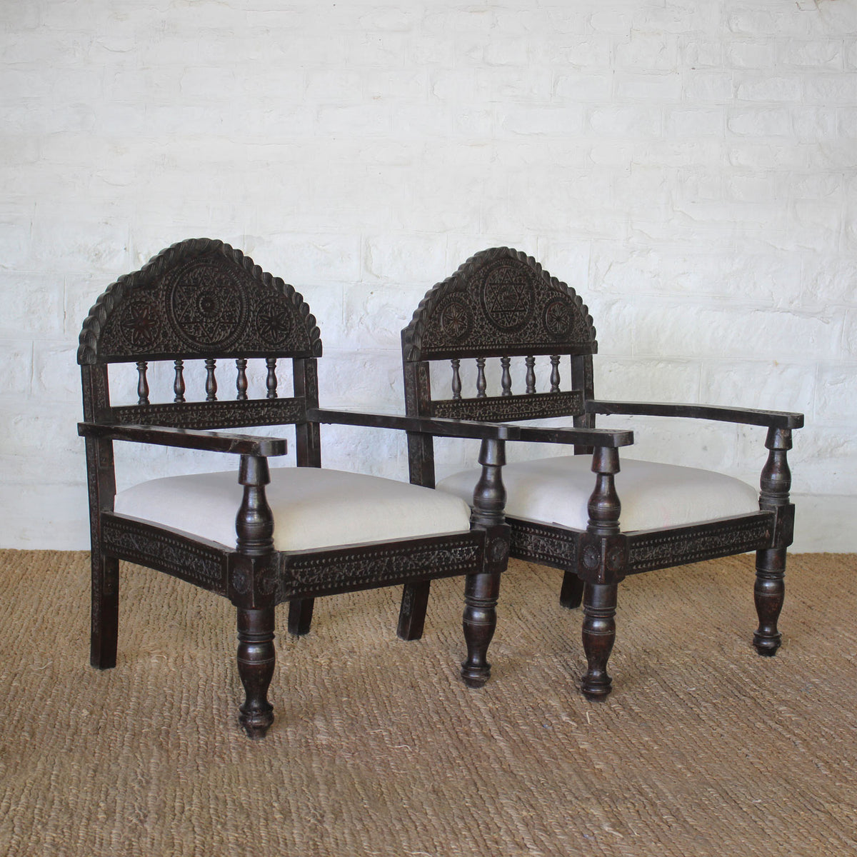 The Himavat Teak Seating Set by John Robshaw, featuring two dark wood armchairs with detailed joinery, hand-carved backrests, and white upholstered seats, is arranged side by side on a woven rug against a white brick wall.