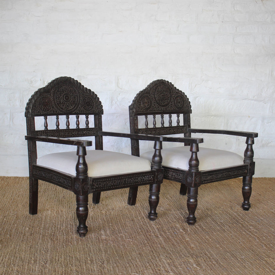 The Himavat Teak Seating Set by John Robshaw, featuring two dark wood armchairs with detailed joinery, hand-carved backrests, and white upholstered seats, is arranged side by side on a woven rug against a white brick wall.