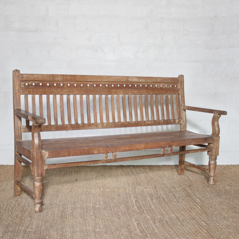 The Mayank Teak Bench by John Robshaw, featuring a slatted back and seat, straight armrests, turned legs, and subtle carved cutout details, is set against a white brick wall on a woven rug.