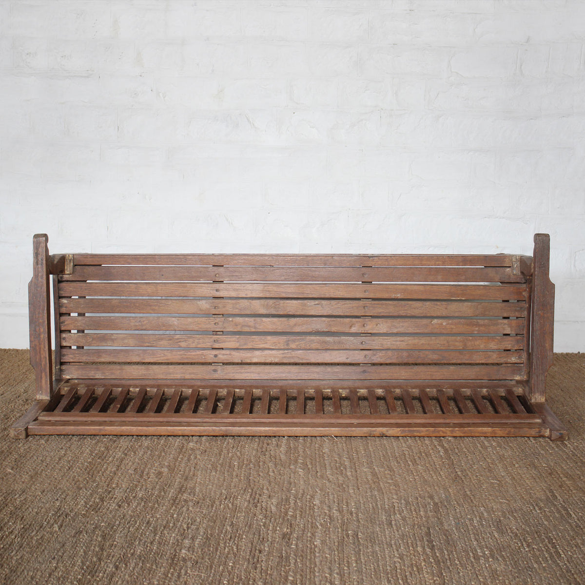 The Mayank Teak Bench by John Robshaw, featuring a slatted back and carved cutout details, is placed on a woven floor mat against a white brick wall.
