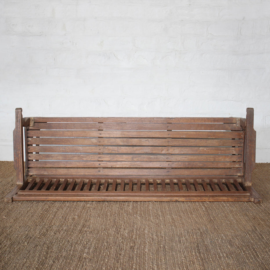 The Mayank Teak Bench by John Robshaw, featuring a slatted back and carved cutout details, is placed on a woven floor mat against a white brick wall.