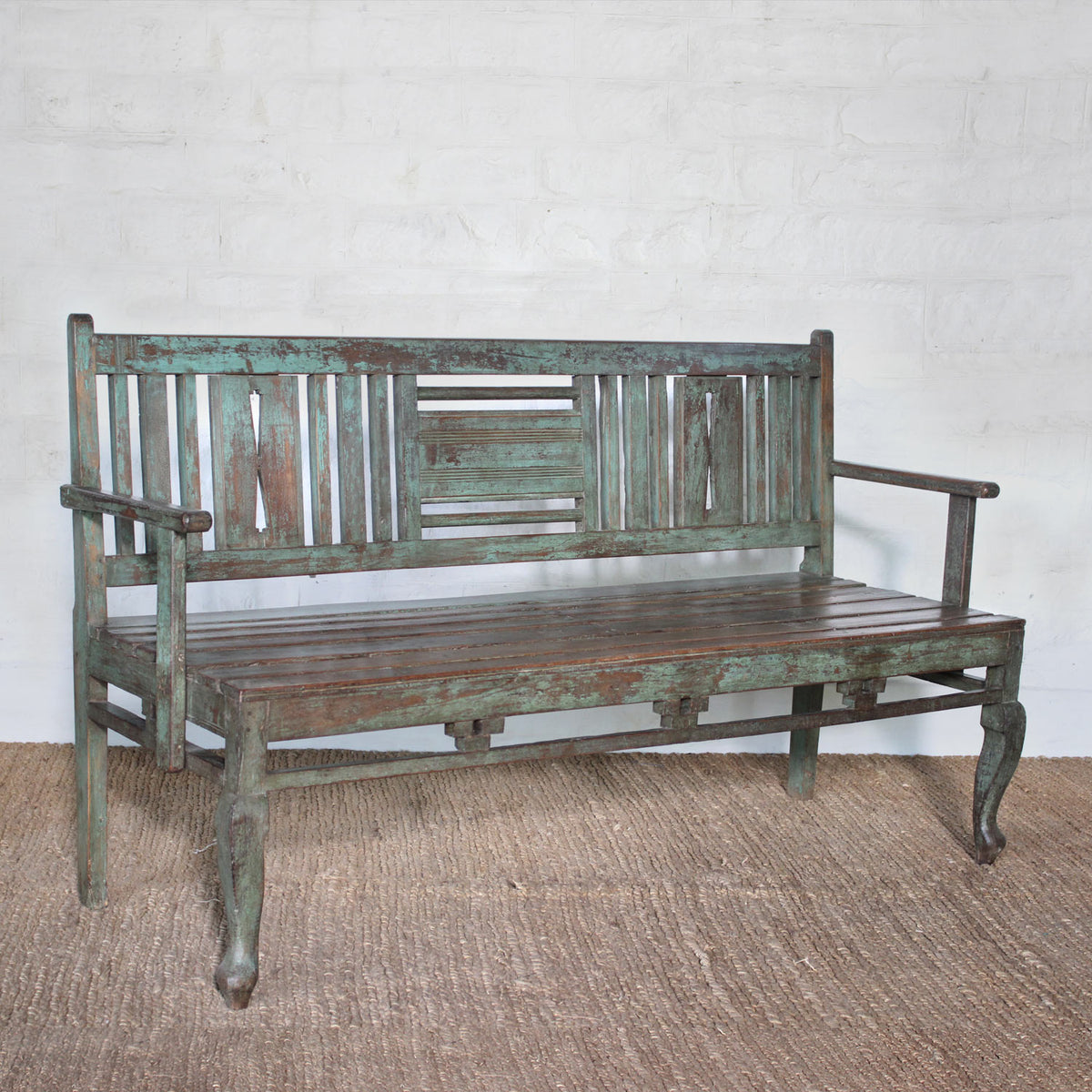 The Pavan Teak Bench by John Robshaw showcases rustic character with its weathered, distressed green finish, slatted backrest, and gracefully curved front legs—perfectly placed on a woven rug against a white brick wall.