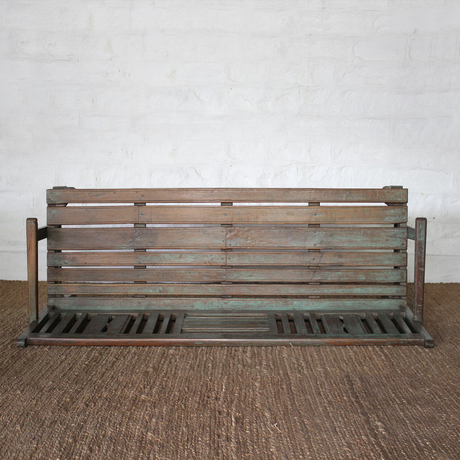 The Pavan Teak Bench by John Robshaw, featuring a weathered finish and rustic character, is set against a white brick wall and positioned on a brown woven rug.