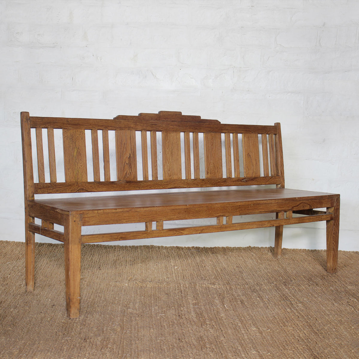The Sarang Teak Bench by John Robshaw, featuring a handcrafted solid teak frame with a slatted backrest and straight legs, sits on a woven rug against a white brick wall.