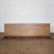 The Sarang Teak Bench by John Robshaw, featuring handcrafted slatted teak seating and a solid backrest, adds the timeless charm of vintage furniture to any space when placed atop a textured brown carpet against a white brick wall.