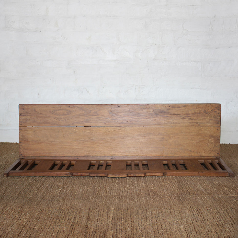 The Sarang Teak Bench by John Robshaw, featuring handcrafted slatted teak seating and a solid backrest, adds the timeless charm of vintage furniture to any space when placed atop a textured brown carpet against a white brick wall.