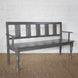 The Varun Teak Bench by John Robshaw, featuring a dark wooden finish with vertical slats on the backrest, armrests, and turned front legs, sits atop a woven rug against a white brick wall, exemplifying the timeless charm of handcrafted vintage furniture.