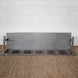 The Varun Teak Bench by John Robshaw, featuring a handcrafted slatted seat and sturdy backrest, is placed atop a woven rug against a white brick wall.
