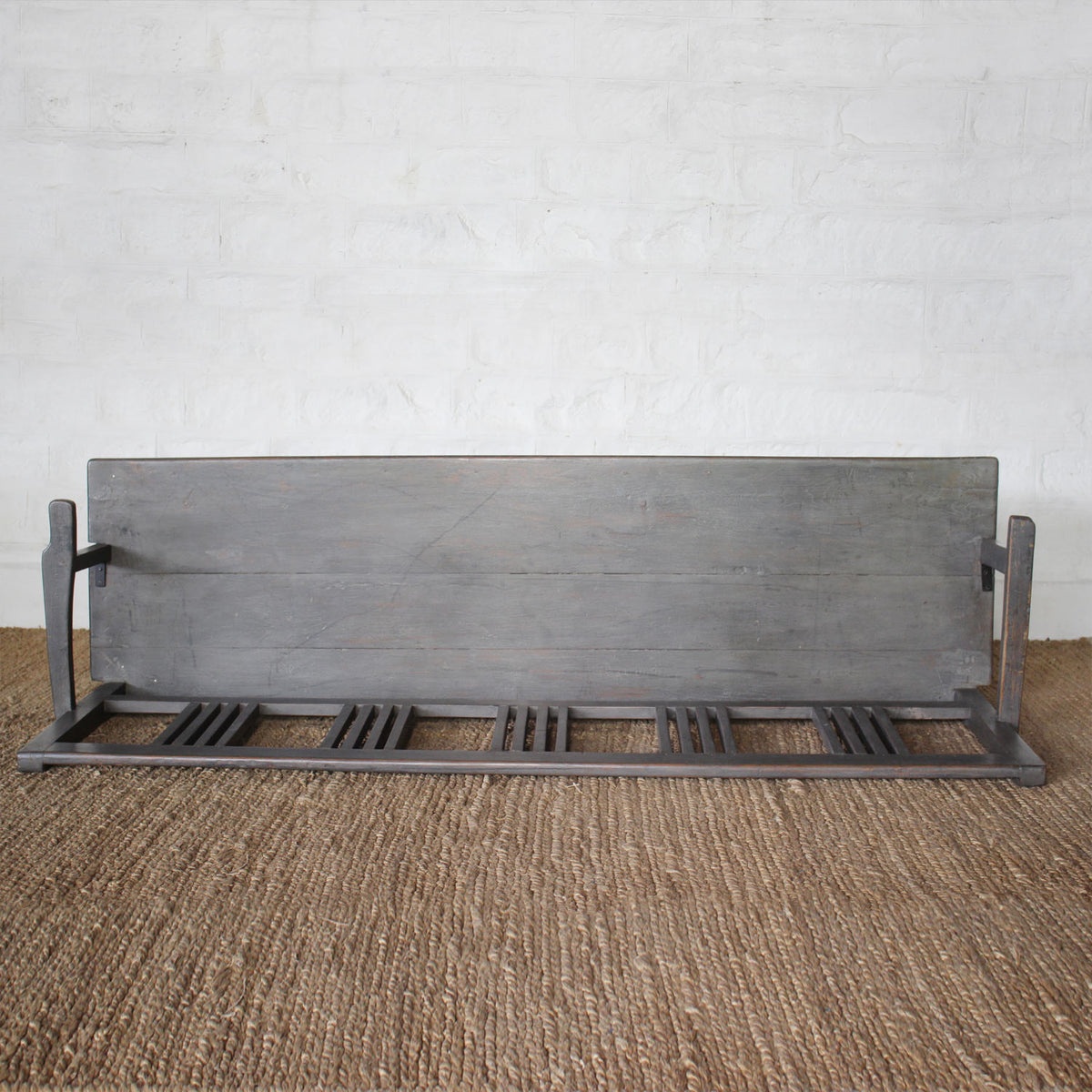The Varun Teak Bench by John Robshaw, featuring a handcrafted slatted seat and sturdy backrest, is placed atop a woven rug against a white brick wall.