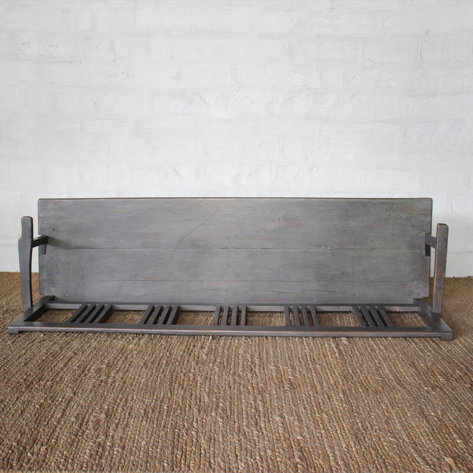 The Varun Teak Bench by John Robshaw, featuring a handcrafted slatted seat and sturdy backrest, is placed atop a woven rug against a white brick wall.