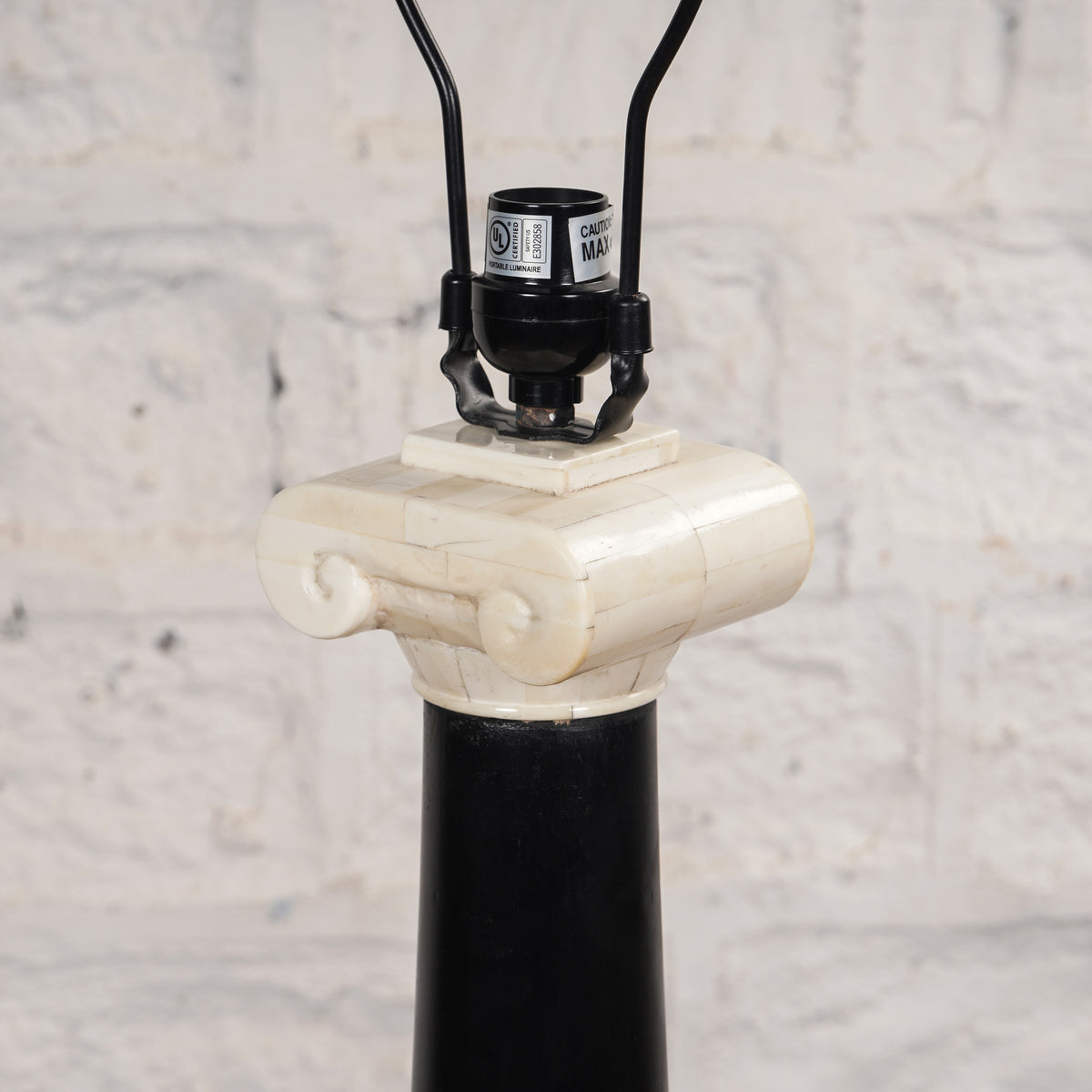 Close-up of the Roshni Bone Inlay Lamp by John Robshaw, featuring a design inspired by a classical column with a black base and a white Ionic capital, evoking the elegance of a Colonial Indian lamp, set against a light-colored textured wall.