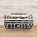 The Rajata Bone Inlay Box by John Robshaw is a rectangular wooden box featuring black and white floral and geometric bone inlay patterns, crafted from Indian teak and displayed on a wooden surface against a light brick wall background.