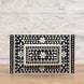 The Rajata Bone Inlay Box by John Robshaw, featuring a rectangular shape and intricate white floral and geometric patterns on a black base, is displayed atop an Indian teak wooden surface with a light brick wall in the background.