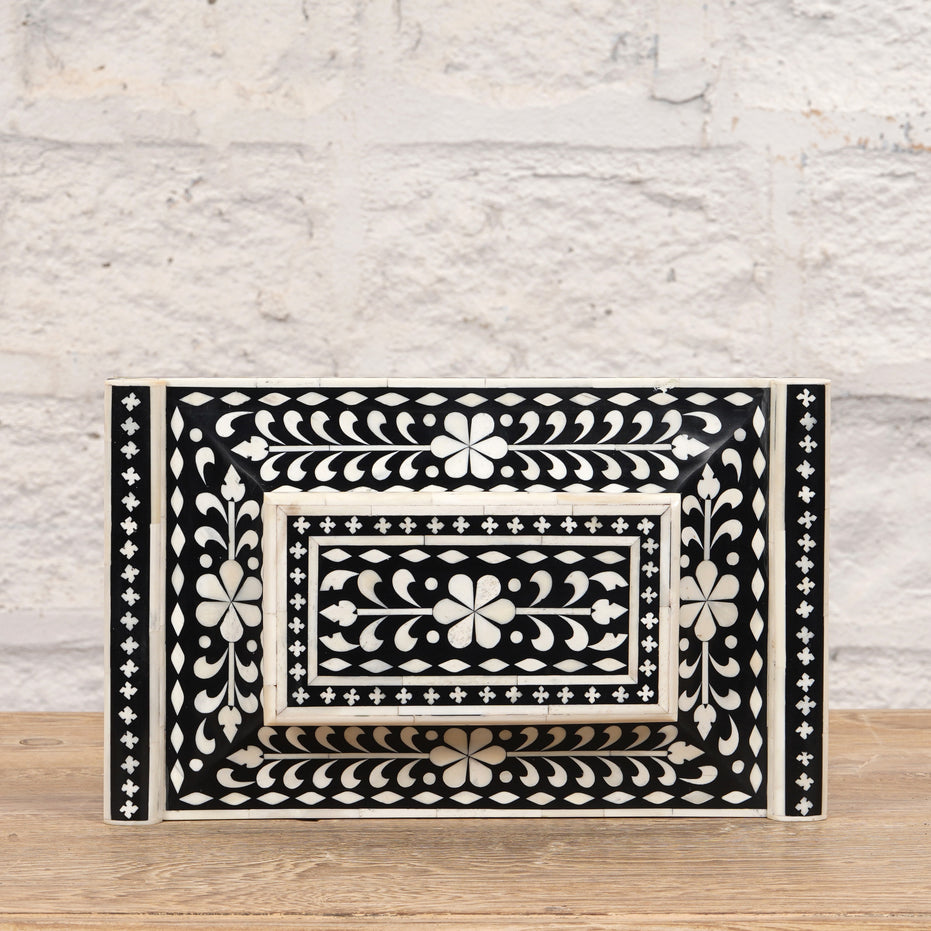 The Rajata Bone Inlay Box by John Robshaw, featuring a rectangular shape and intricate white floral and geometric patterns on a black base, is displayed atop an Indian teak wooden surface with a light brick wall in the background.