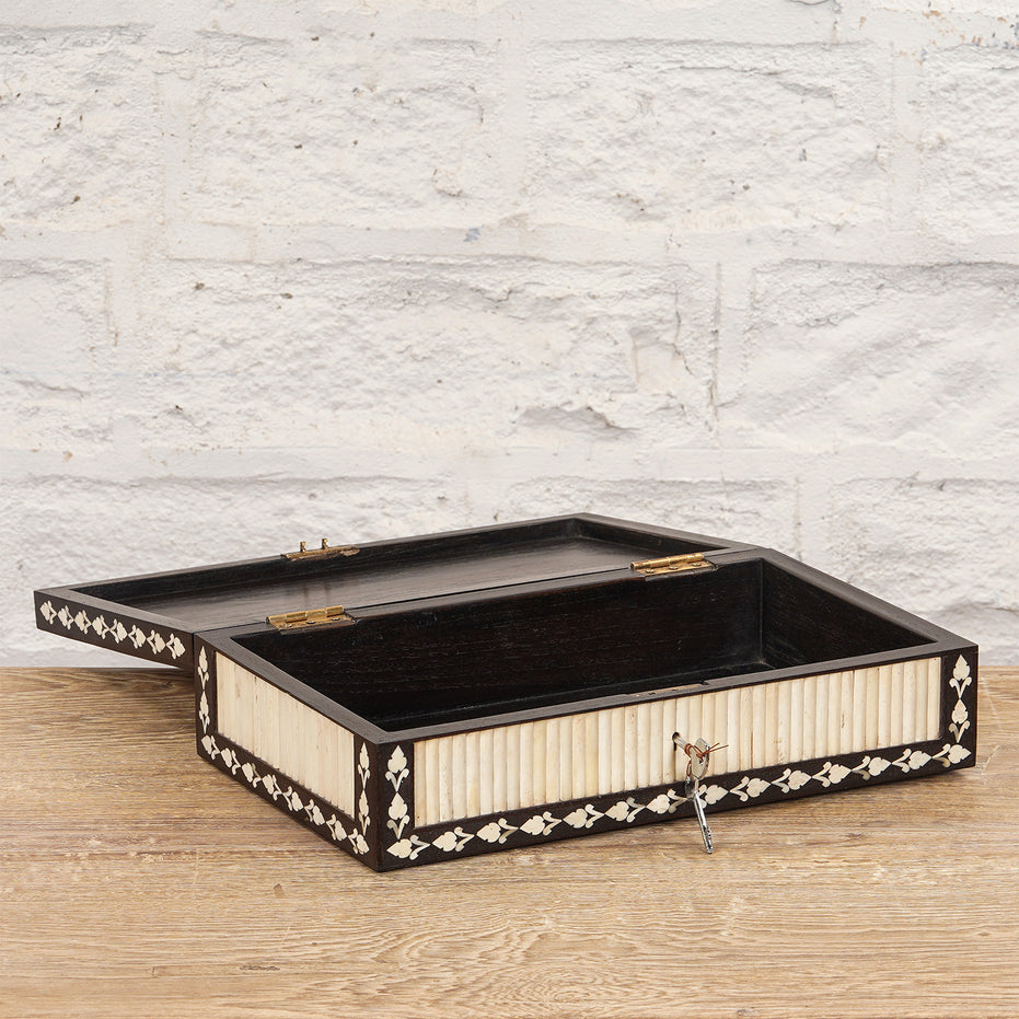 The Charita Bone Inlay Box by John Robshaw features a rectangular teak design with a subtle striped white bone inlay pattern. Shown with its lid open and a metal key in the lock, the box rests on a wooden surface set against a white brick wall.