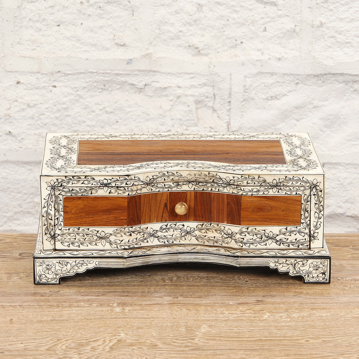 The Harini Bone Inlay Box by John Robshaw is a rectangular handcrafted teak box featuring an intricate floral inlay design, a single small front drawer with a brass knob, and sits gracefully on a wooden surface against a white brick wall—this vintage Indian-inspired piece infuses any space with timeless charm.