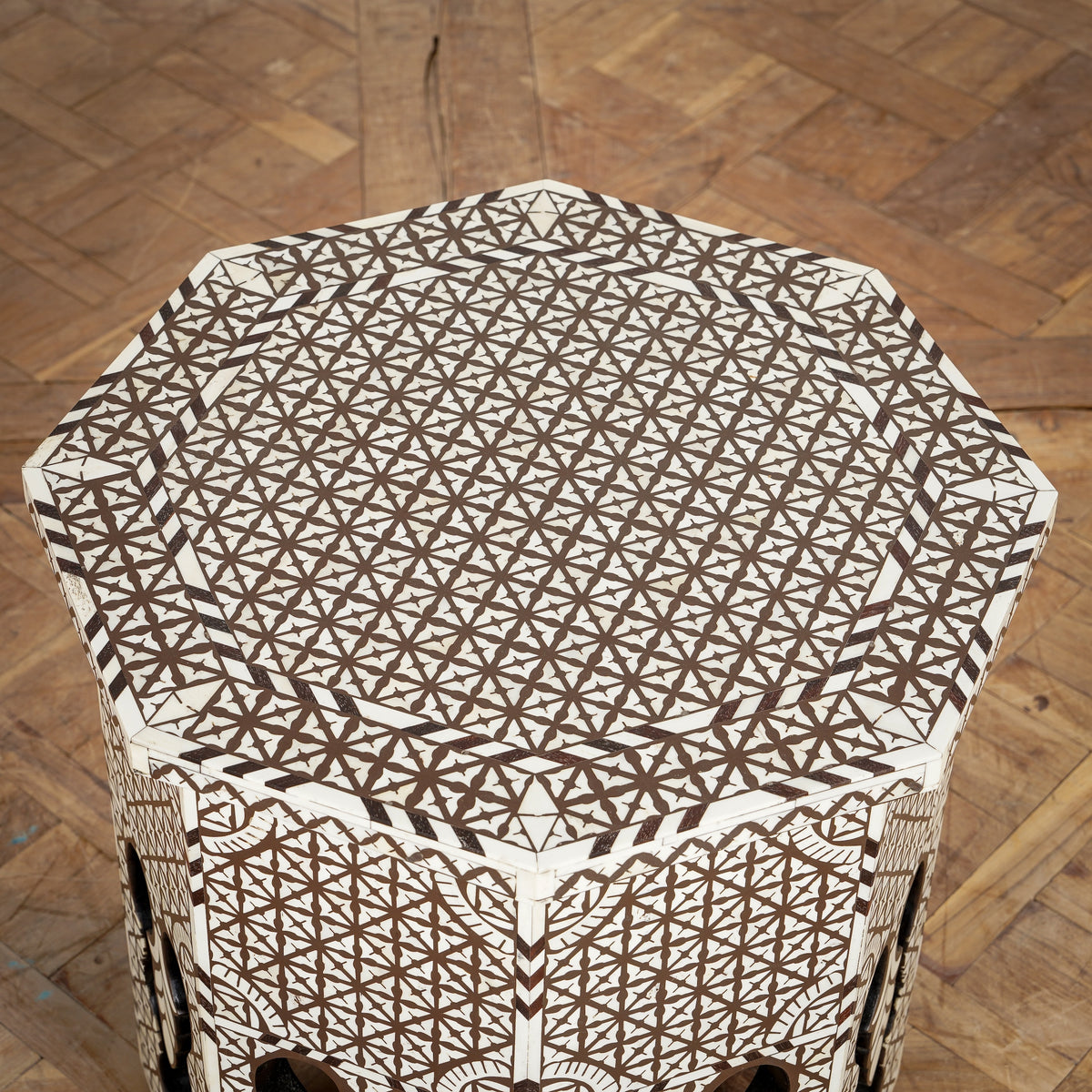 The Chandni Bone Inlay Table by John Robshaw features an octagonal wooden design adorned with intricate geometric patterns in brown and ivory, set atop a parquet wood floor—a striking piece of bone inlay furniture that adds timeless elegance to any space.