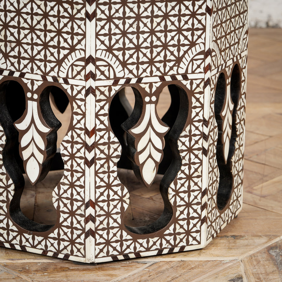 The Chandni Bone Inlay Table by John Robshaw features intricate geometric star and floral patterns delicately inlaid in white, reminiscent of traditional bone inlay craftsmanship, with striking arched cutouts along its sides, and is pictured here on a wooden floor.