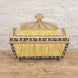 The Suvarna Bone Inlay Box by John Robshaw features a gold and black floral design, ribbed side panels, and a domed lid crowned with a gold finial. Resting on a wooden surface against a white brick wall, this striking piece is reminiscent of an Indian teak box.