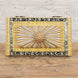 The Suvarna Bone Inlay Box by John Robshaw features a decorative combination of gold, silver, and black accents with a raised floral and sunburst design on the lid, beautifully displayed on a wooden surface against a white brick wall.