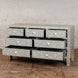 The Vajra Bone Inlay Sideboard by John Robshaw features eight floral-patterned drawers, each partially open, and stands on a wooden floor against a white brick wall. This exquisite piece is both artistic and functional, serving as a true statement in any space.