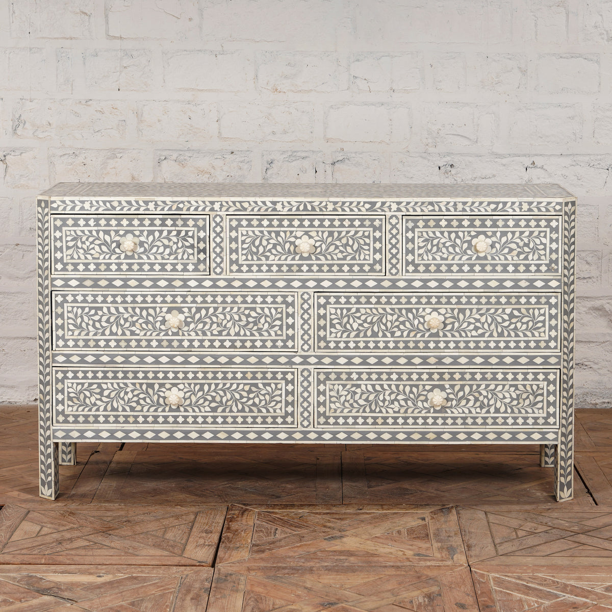 The Vajra Bone Inlay Sideboard by John Robshaw features a striking gray and white pattern with six drawers, adorned with decorative floral and geometric bone inlay, serving as a statement piece on a wooden floor against a white brick wall.