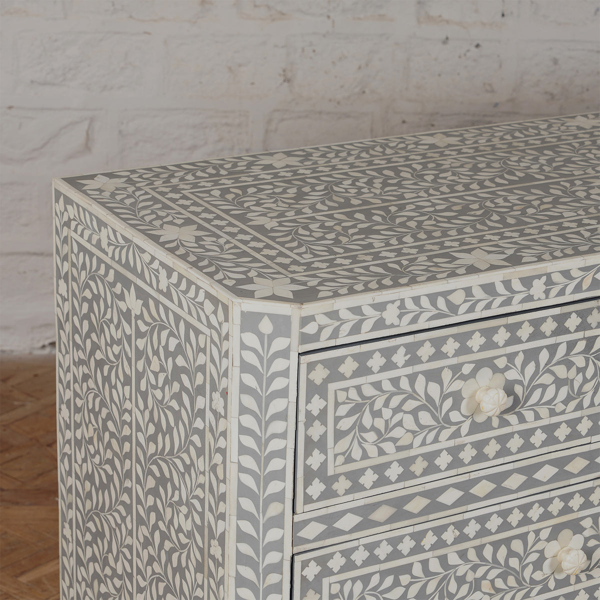 A close-up of the Vajra Bone Inlay Sideboard by John Robshaw showcases its intricate white and gray floral patterns, set against a white brick wall and wooden floor, making it a stunning statement piece for any room.