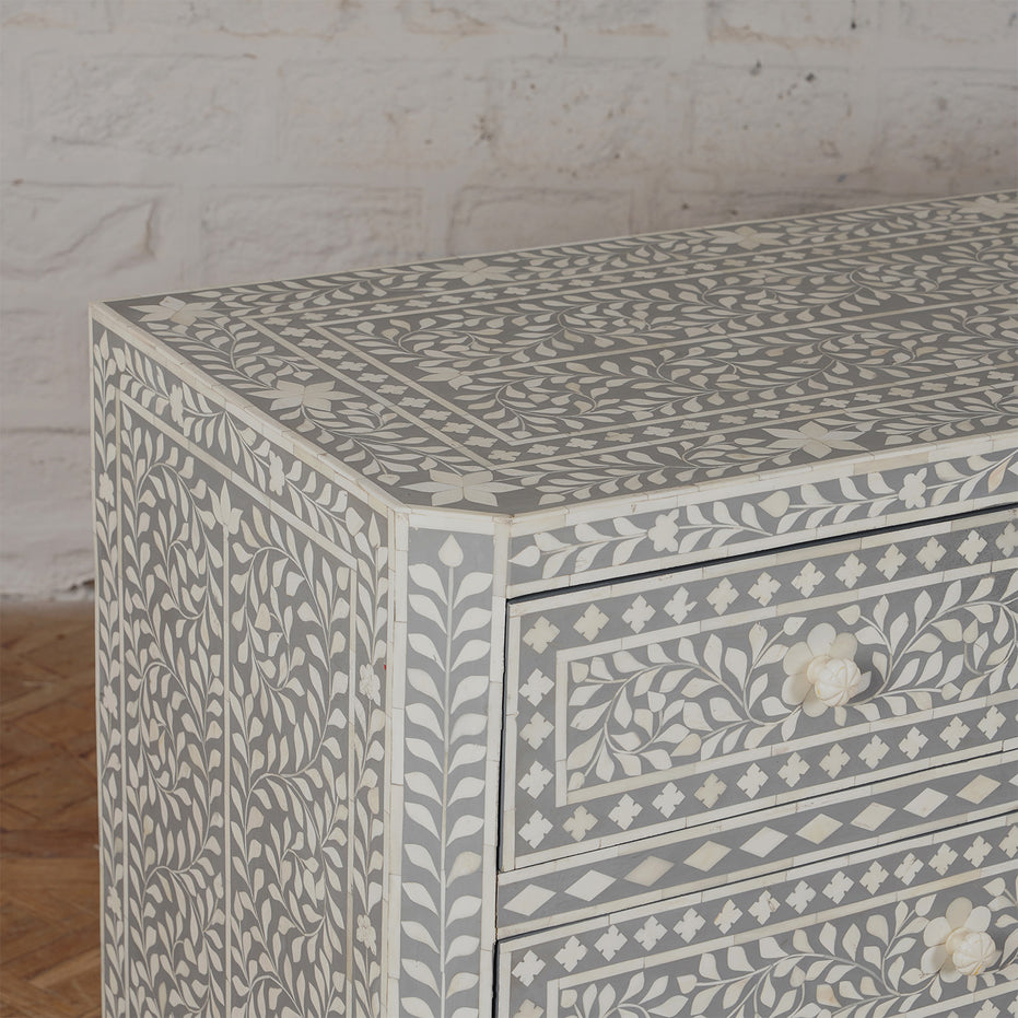 A close-up of the Vajra Bone Inlay Sideboard by John Robshaw showcases its intricate white and gray floral patterns, set against a white brick wall and wooden floor, making it a stunning statement piece for any room.