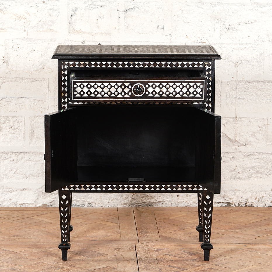 The John Robshaw Ahan Bone Inlay Cabinet showcases intricate black and white geometric patterns, featuring one drawer and an open cabinet door, all standing on four tapered legs against a white brick wall.