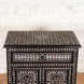 Set against a white brick wall, the Ahan Bone Inlay Cabinet by John Robshaw features intricate black and white geometric inlay patterns on the top, drawer, and doors. This stunning piece showcases refined bone inlay work that adds a touch of elegance to any space.