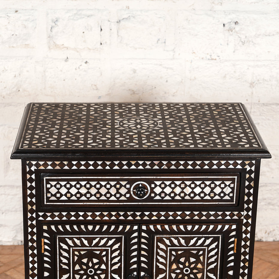 Set against a white brick wall, the Ahan Bone Inlay Cabinet by John Robshaw features intricate black and white geometric inlay patterns on the top, drawer, and doors. This stunning piece showcases refined bone inlay work that adds a touch of elegance to any space.