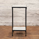 The Veda Side Table by John Robshaw is a small, rectangular two-tier minimalist piece, featuring a black iron frame with light-colored surfaces, shown here standing on a parquet wood floor against a white brick wall.
