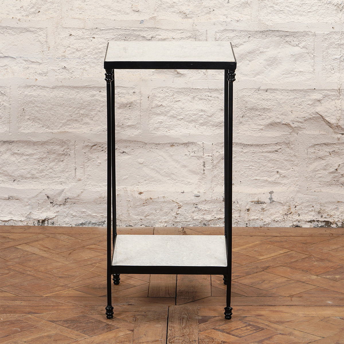 The Veda Side Table by John Robshaw is a small, rectangular two-tier minimalist piece, featuring a black iron frame with light-colored surfaces, shown here standing on a parquet wood floor against a white brick wall.